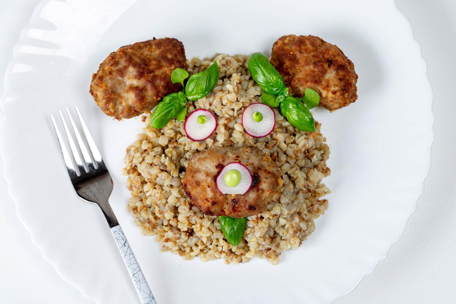 vue de dessus du visage d'un ours fait de bouillie de sarrasin, boulettes de viande, légumes et basilic sur une assiette blanche photo