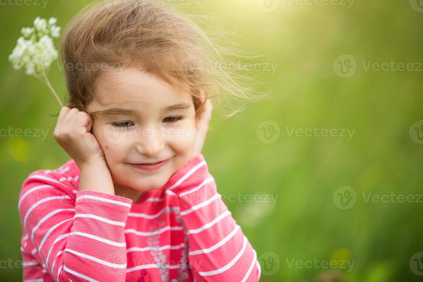 petite fille dans un t-shirt à rayures corail sur fond vert dans un champ tient son visage dans ses mains et sourit sournoisement. journée des enfants, enfant heureux, protection de l'environnement et de la nature, insectifuge photo