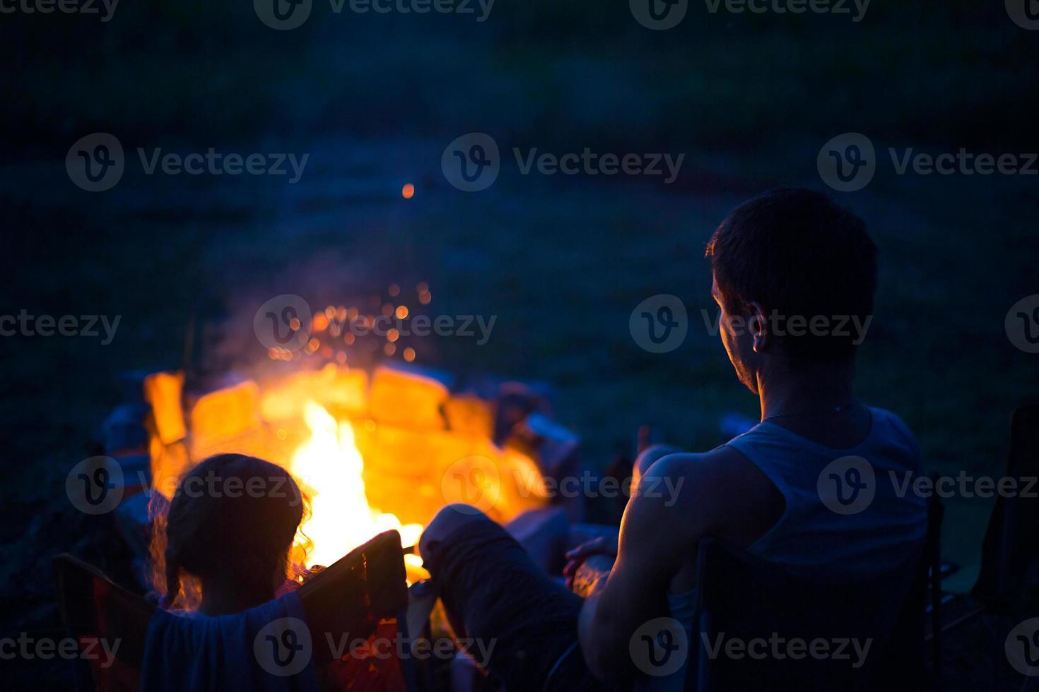 papa et sa fille s'assoient la nuit au coin du feu en plein air en été dans la nature. voyage de camping en famille, rassemblements autour du feu de camp. fête des pères, barbecue. lanterne et tente de camping photo