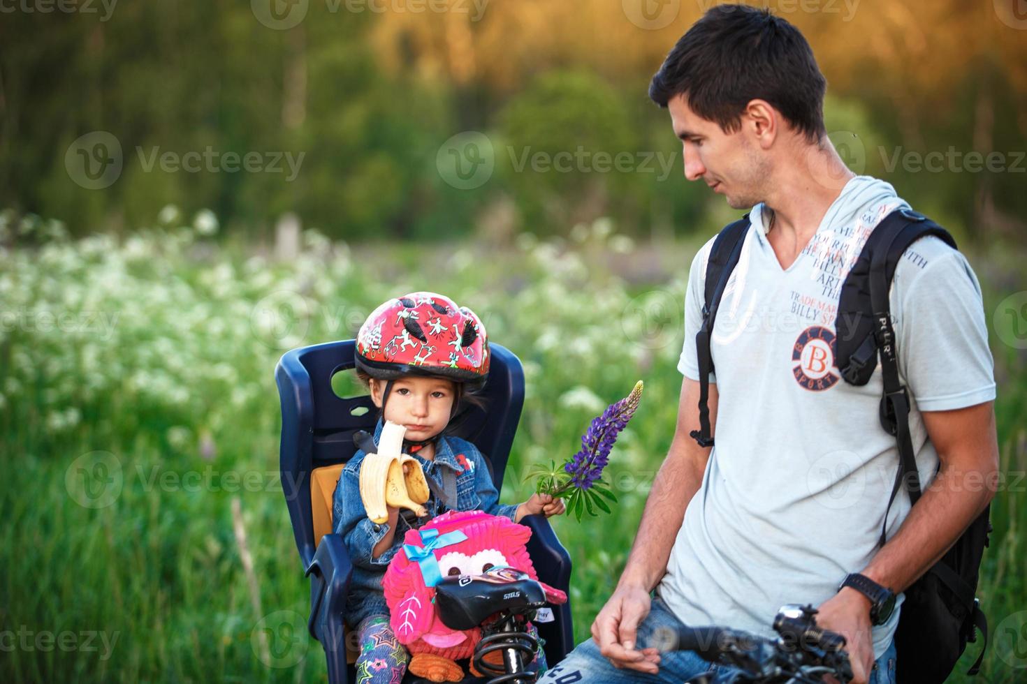 père avec une petite fille dans un siège de vélo pour enfants sur un vélo adulte. une fille dans un casque de protection, avec une banane et un sac à dos. balade sportive familiale, sécurité. Kaluga, Russie, 30 mai 2018 photo