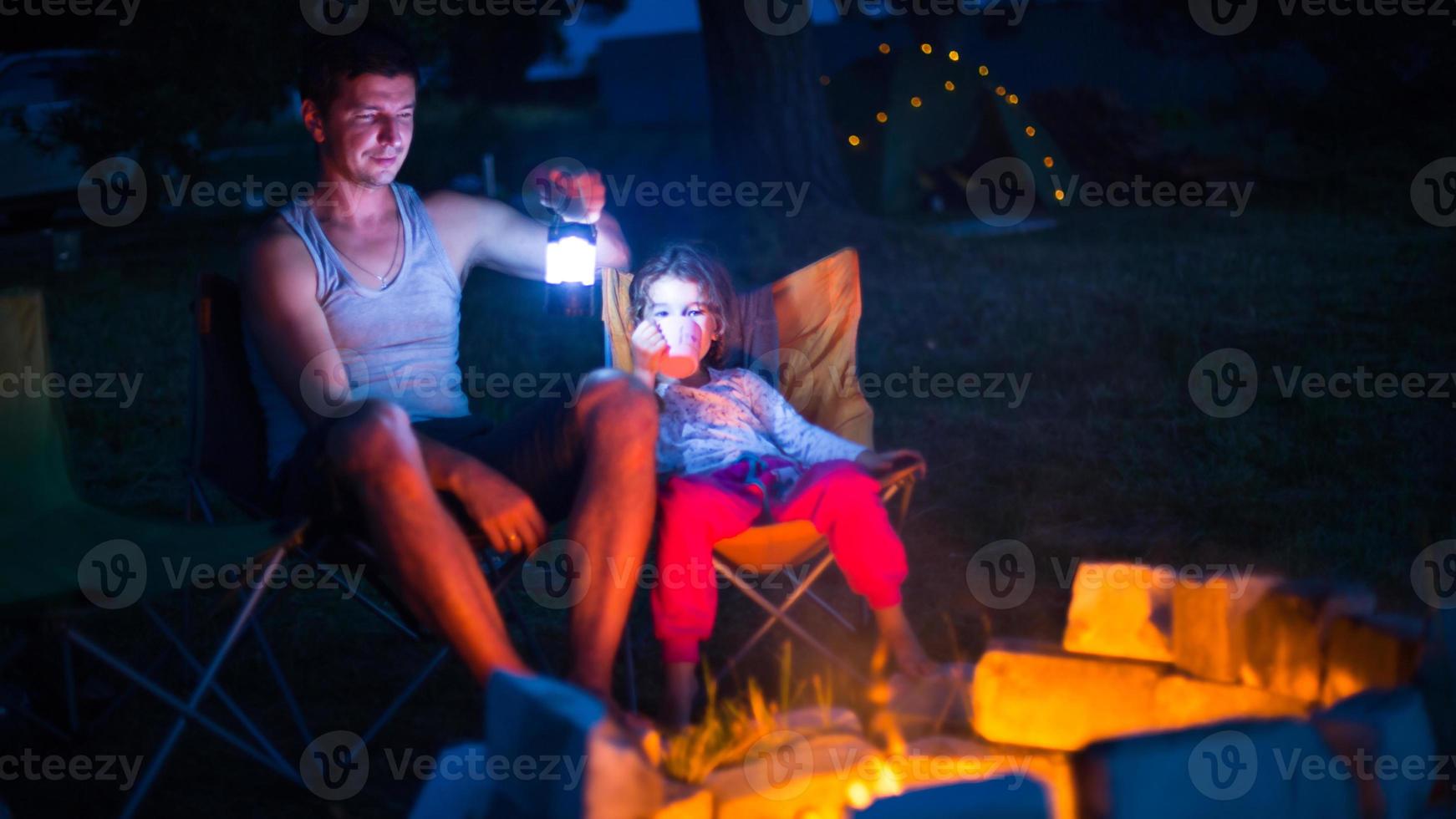 papa et sa fille s'assoient la nuit au coin du feu en plein air en été dans la nature. voyage de camping en famille, rassemblements autour du feu de camp. fête des pères, barbecue. lanterne et tente de camping photo