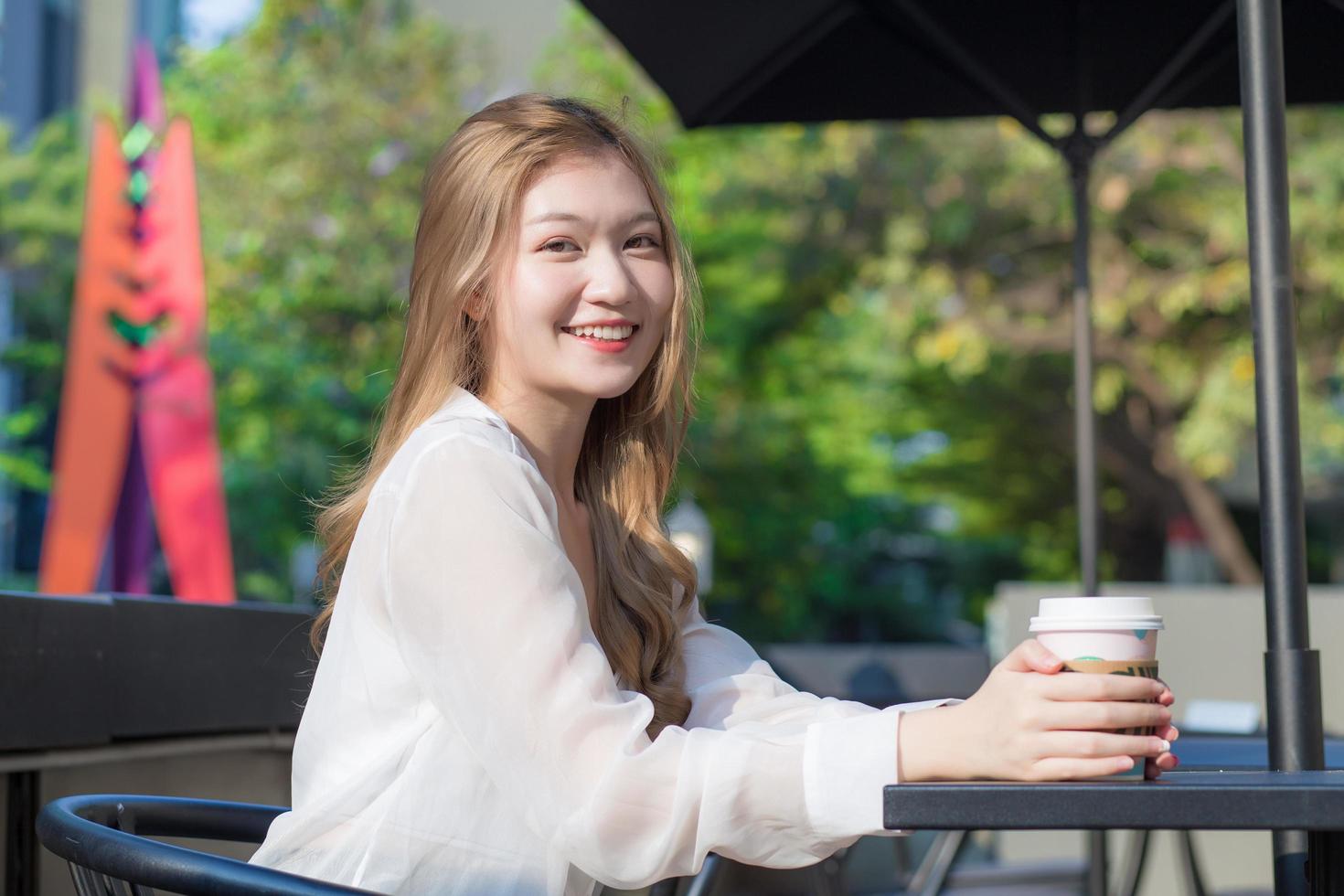 jeune belle femme asiatique qui porte un costume aux cheveux bronze est assis sourire sur une chaise dans un café à l'extérieur tout en tenant une tasse de café dans sa main un matin ensoleillé. photo