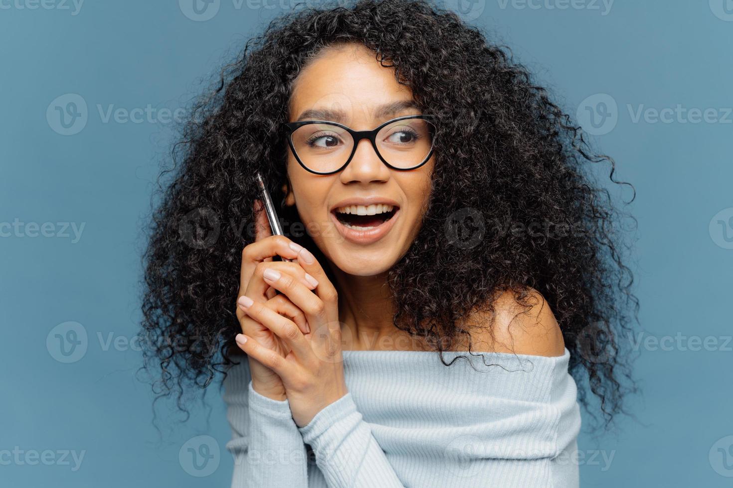 photo de tête d'une jeune femme afro-américaine séduisante tenant un téléphone portable moderne près de l'oreille, a une belle conversation, concentrée de côté, remarque quelque chose de drôle et d'inattendu, isolée sur fond bleu