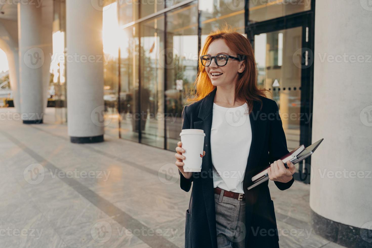 le modèle féminin tient un ordinateur portable à pavé tactile et une tasse de café à emporter pose en plein air pendant la journée ensoleillée photo