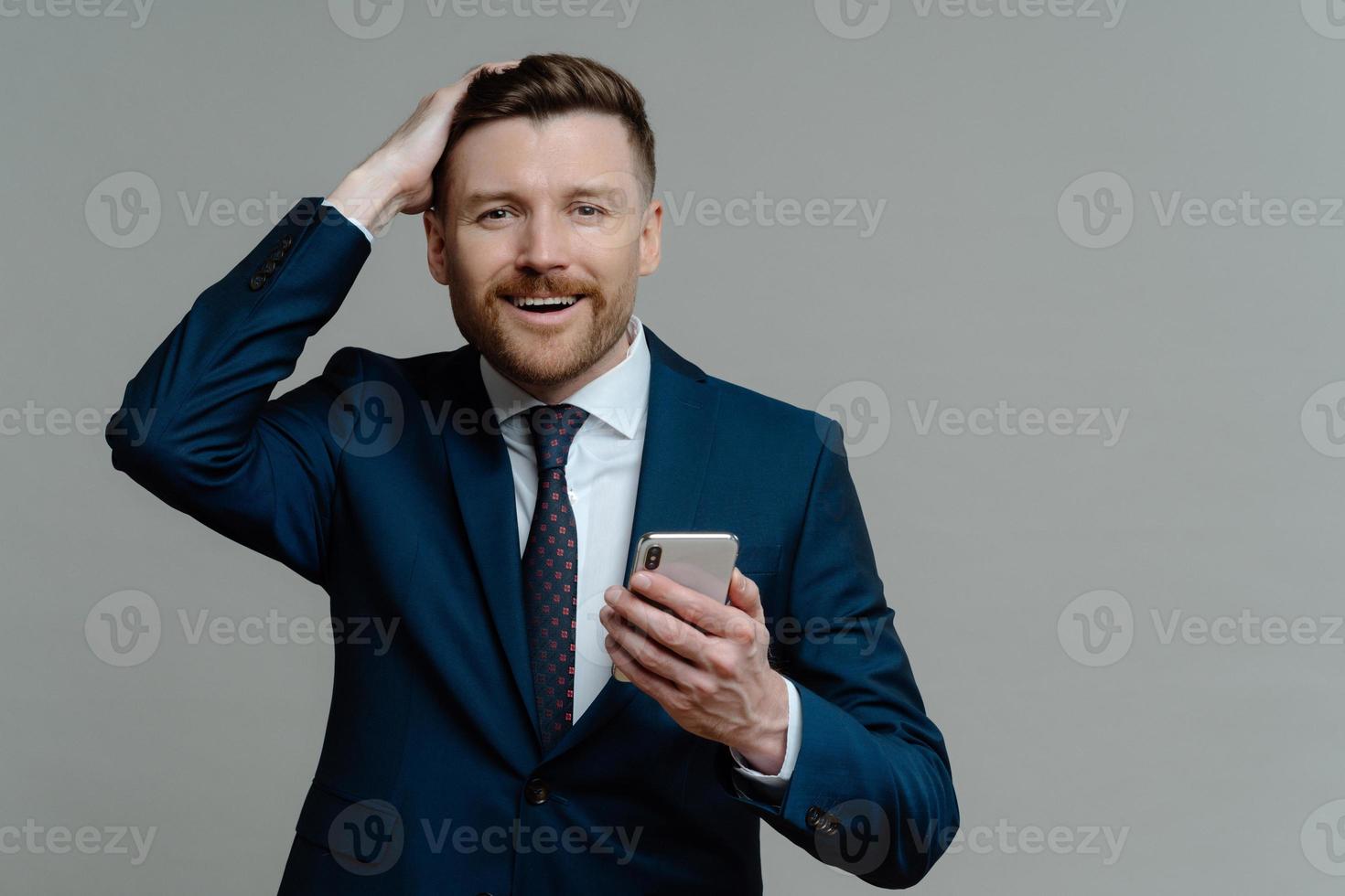 photo horizontale d'un bel homme d'affaires barbu garde la main sur la tête sourit joyeusement tient le smartphone utilise la plate-forme pour la banque habillé formellement isolé sur fond gris. administrateur de société