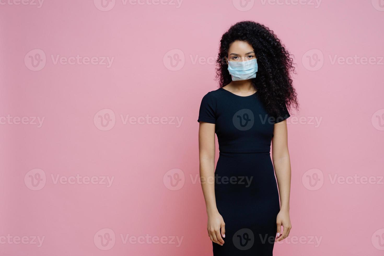 une femme afro-américaine sérieuse porte un masque médical jetable sur le visage, étant en auto-isolement pendant la quarantaine, reste seule à la maison, présente des symptômes de coronavirus, se dresse sur fond rose photo