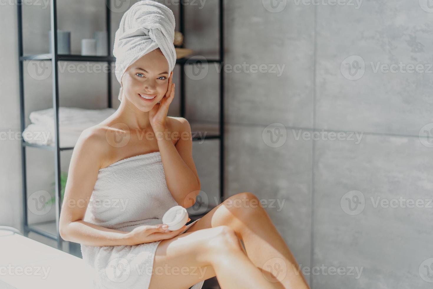 photo d'une jeune femme européenne souriante touche la joue, applique une crème hydratante sur le visage, a une expression heureuse, enveloppée dans une serviette de bain douce, pose dans la salle de bain. concept de routine d'hygiène beauté