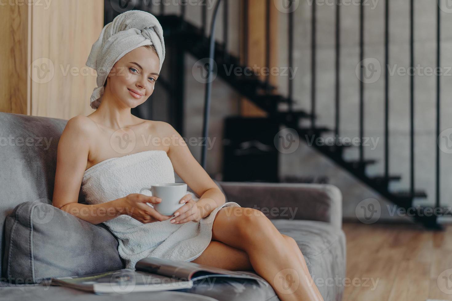 photo horizontale d'une jeune femme européenne heureuse sourit doucement pose enveloppée dans une serviette boit du café pose sur un canapé dans le salon lit un magazine, applique une crème de beauté sur le visage après avoir pris une douche