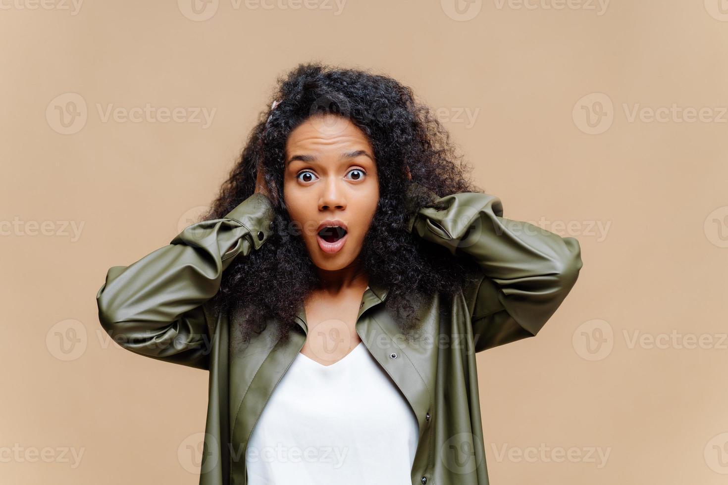 une femme afro-américaine impressionnée garde les deux mains sur la tête, ouvre largement la bouche, vêtue d'un t-shirt blanc et d'une chemise en cuir, émerveillée par quelque chose, isolée sur fond marron. omg concept photo