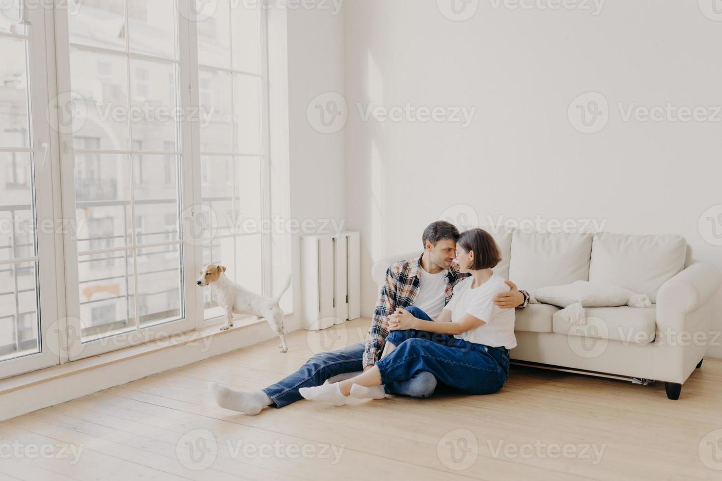 vue horizontale d'un couple familial heureux et affectueux vêtu de vêtements décontractés, s'embrassent et s'expriment l'amour, posent sur le sol près du canapé dans un appartement moderne, leur animal de compagnie regarde à travers une grande fenêtre photo