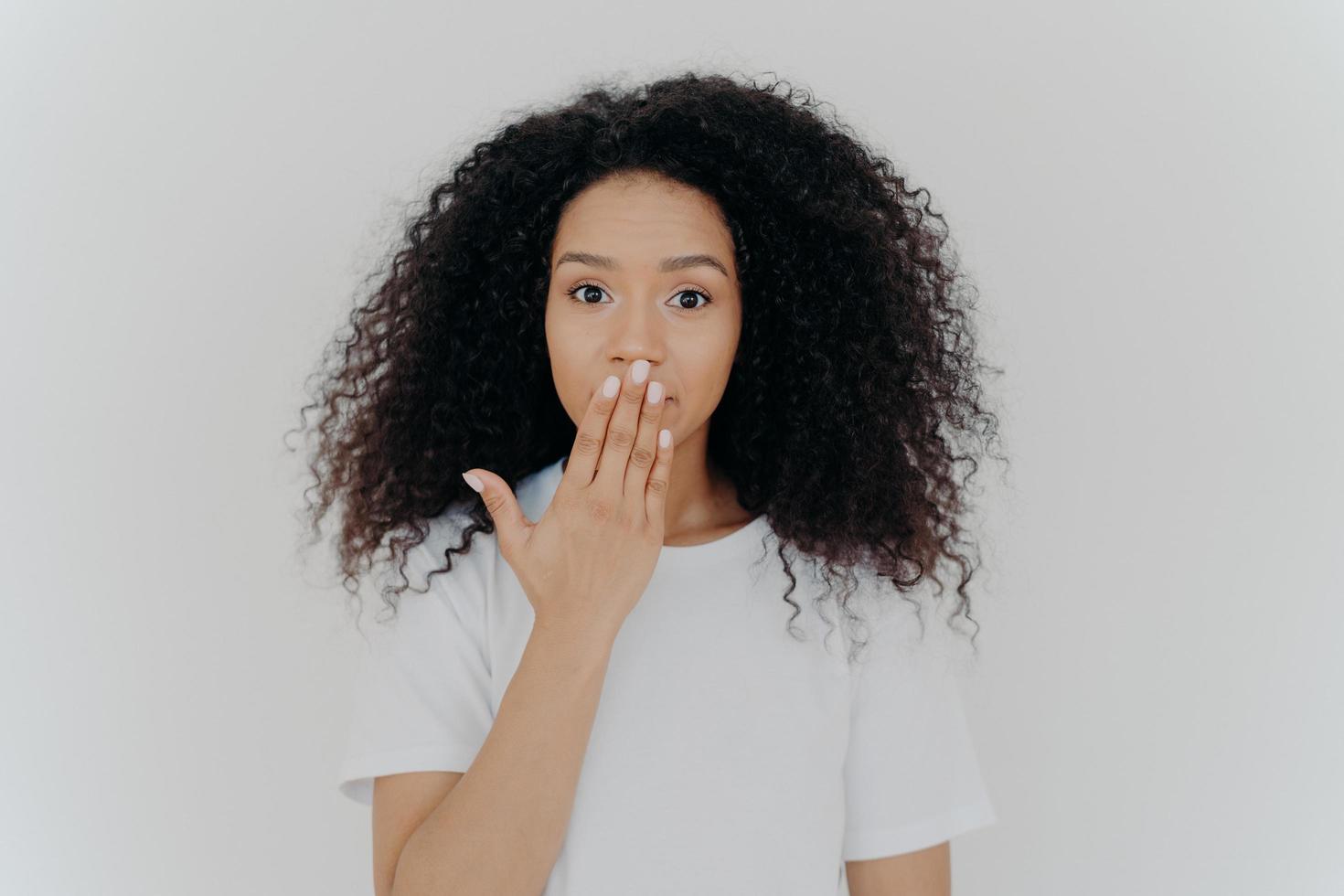 photo de tête d'une jeune femme bouclée impressionnée couvre la bouche avec la paume, essaie d'être sans voix, a une coiffure bouclée, une manucure et un maquillage minimal, porte un t-shirt blanc, pose à l'intérieur. omg, quels potins