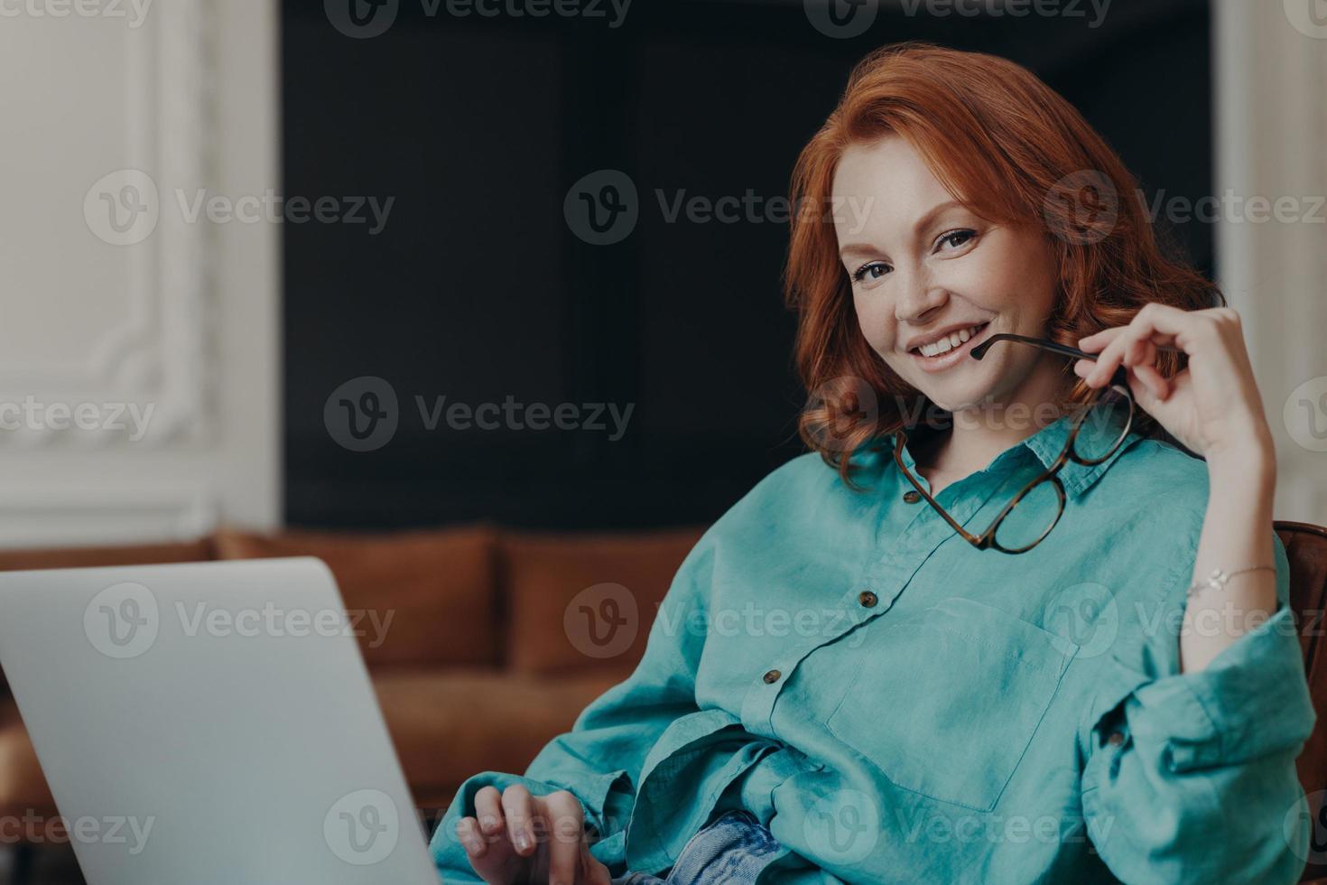 Une belle jeune femme indépendante positive travaille à distance sur un ordinateur portable, connectée à Internet sans fil gratuit à la maison, tient des lunettes, vêtue d'une chemise, sourit joyeusement, est de bonne humeur photo