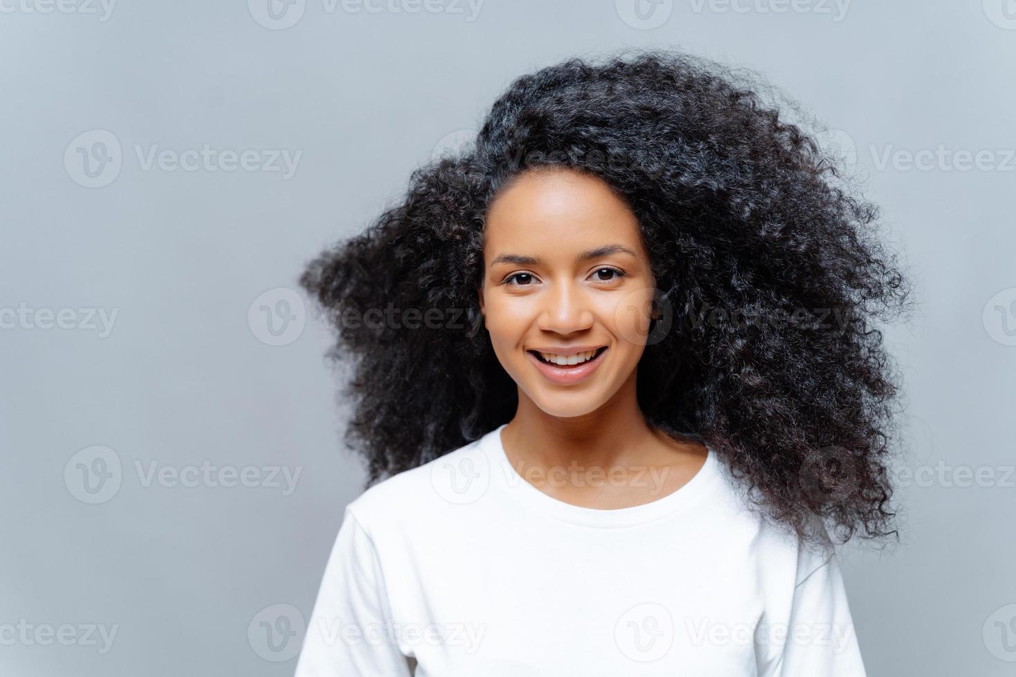 femme bouclée positive à la beauté naturelle, vêtue d'un t-shirt décontracté blanc, a une expression heureuse, regarde directement la caméra, pose sur fond gris. joyeuse adolescente exprime de bonnes émotions photo