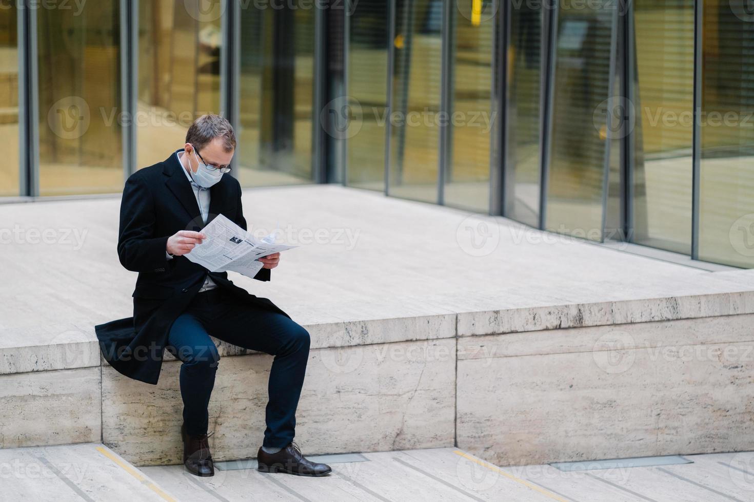 photo extérieure horizontale d'un homme d'affaires ou d'un entrepreneur pose à l'extérieur dans la rue, lit le journal avec un regard attentif, porte un masque médical pour se protéger contre l'infection ou le coronavirus. protection de soi