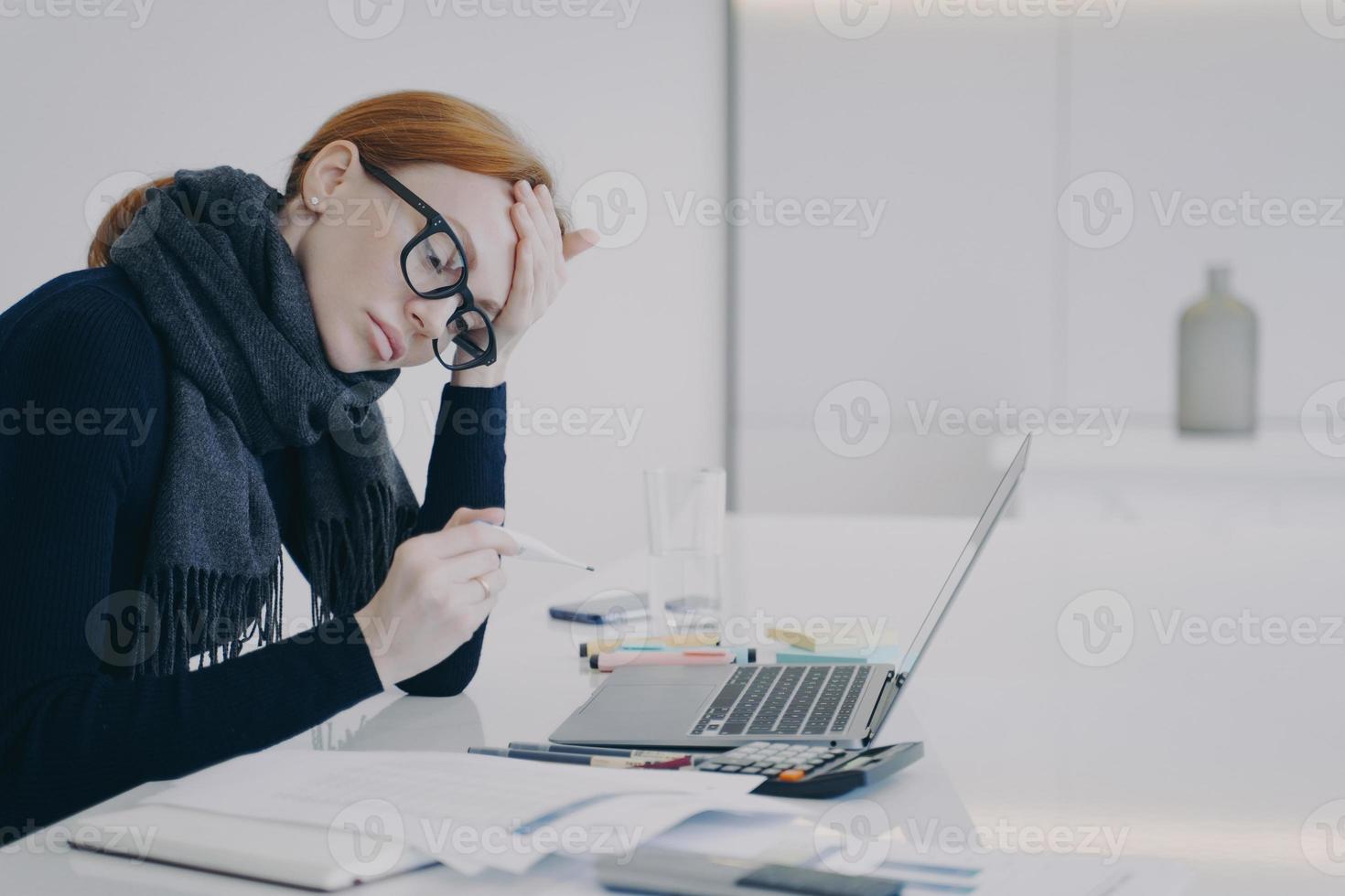 secrétaire est malade. jeune femme blanche au bureau tenant un thermomètre mais faisant son travail. photo