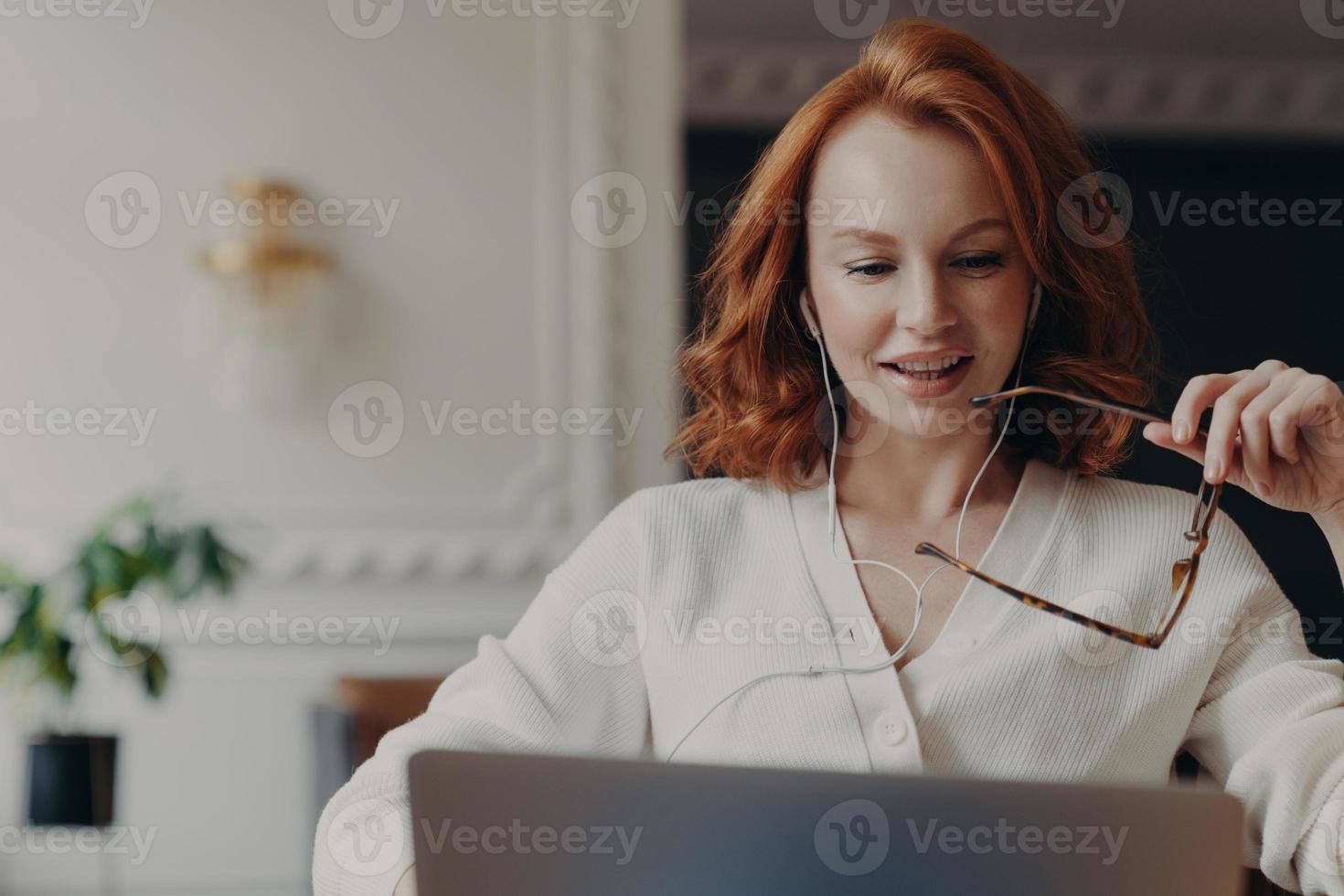 une étudiante rousse heureuse regarde un webinaire de formation, a une vidéoconférence en ligne, utilise des écouteurs, travaille sur un projet d'entreprise, garde des lunettes transparentes à la main. travail à distance via internet. photo