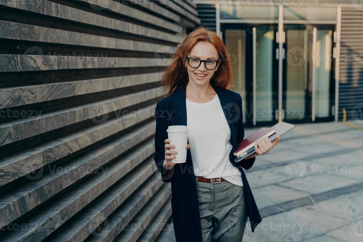 femme d'affaires réussie au gingembre debout à l'extérieur près du bureau avec ordinateur portable, ordinateur portable et café photo