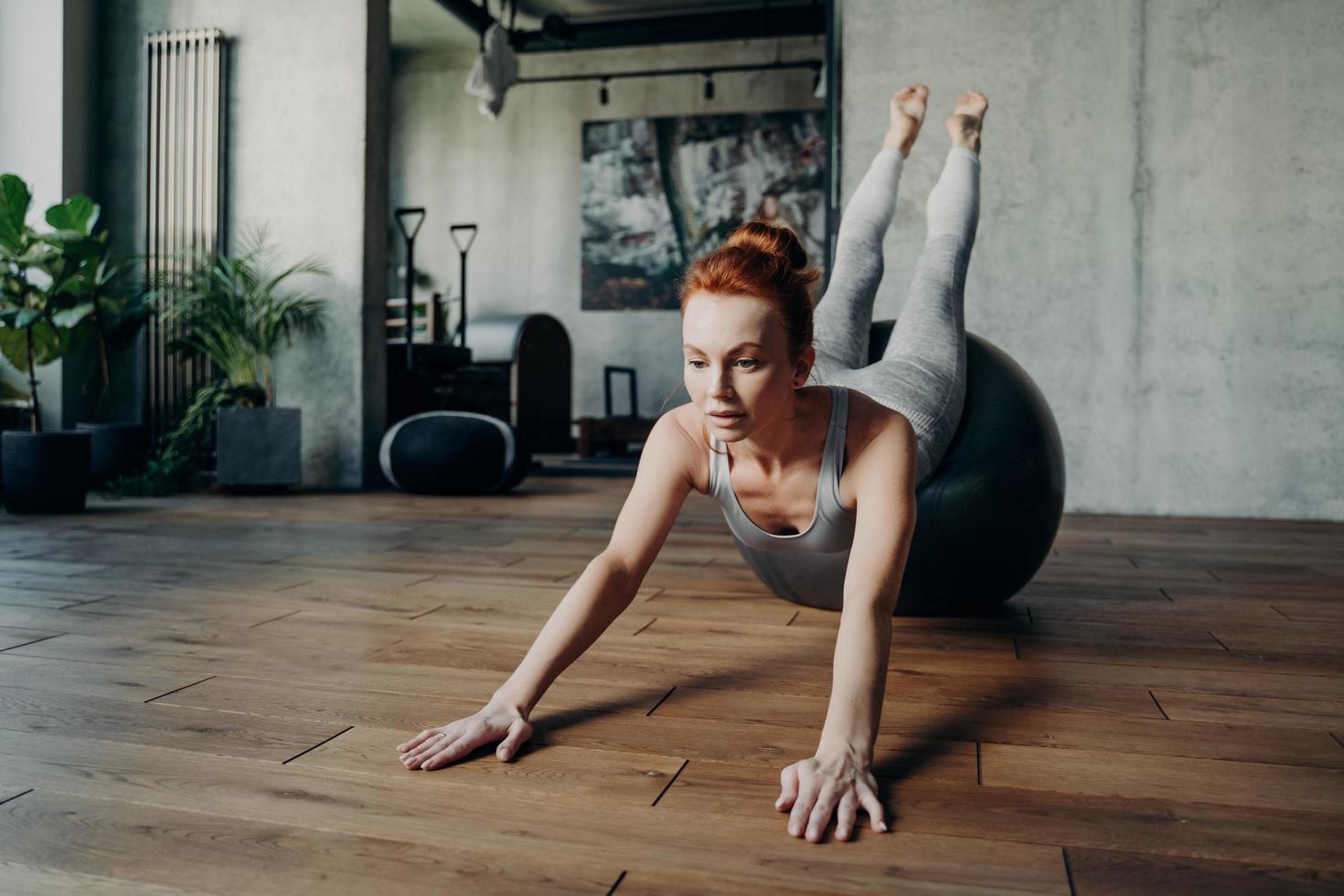 Dame mince athlétique aux cheveux rouges effectuant des exercices d'étirement sur un grand fitball argenté pendant l'entraînement de pilates photo