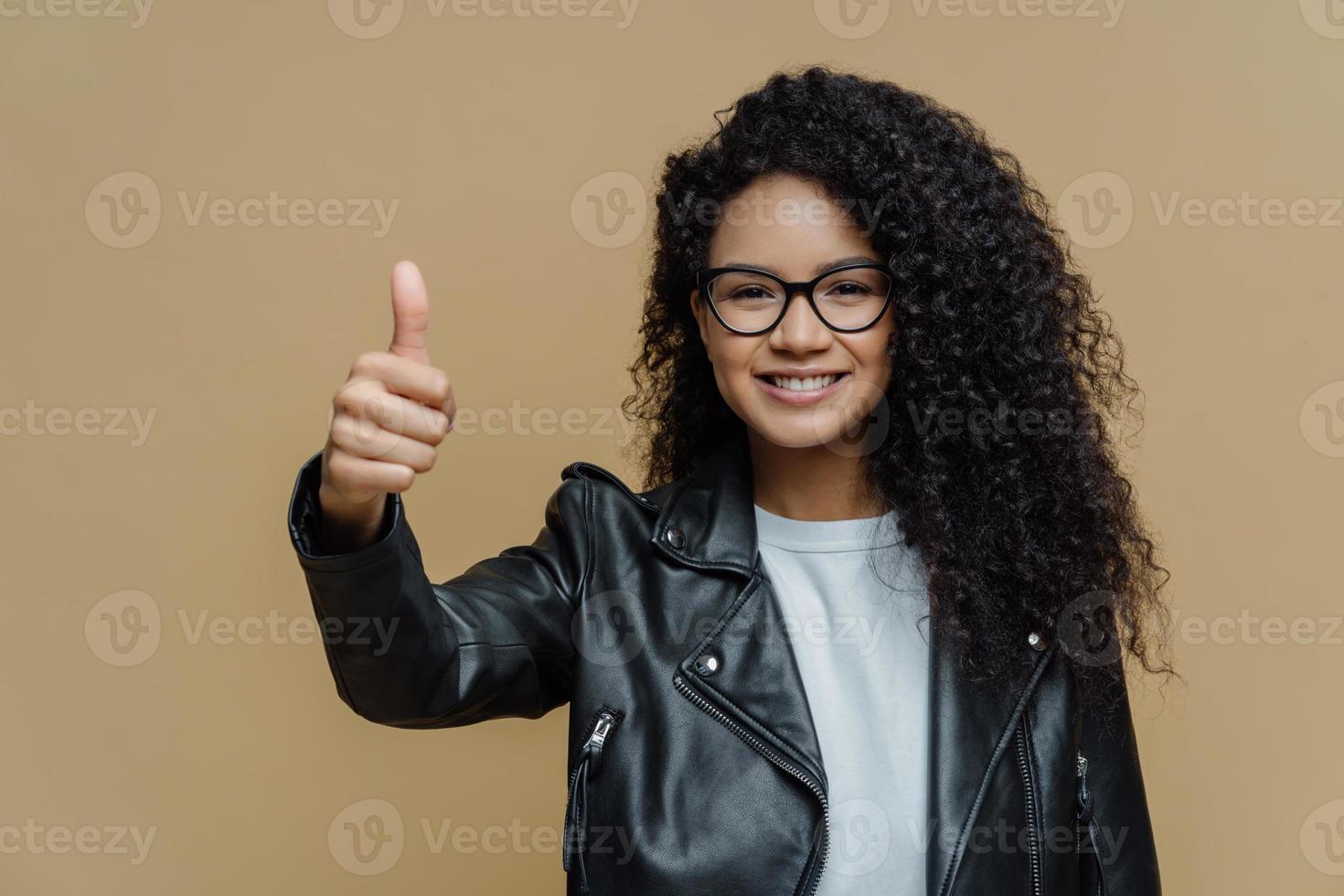 Je suis totalement d'accord avec vous. une femme aux cheveux bouclés ravie montre le pouce vers le haut, évalue un excellent produit, recommande une offre promotionnelle, porte des lunettes et une veste en cuir, isolée sur fond beige, approuve la meilleure offre photo