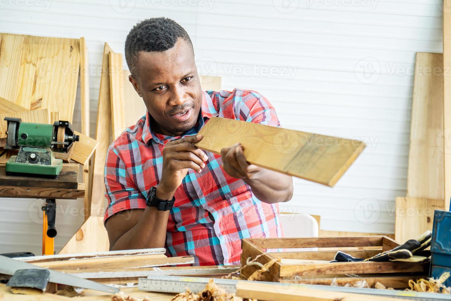 jeune charpentier afro-américain regardant et choisissant du bois et utilisant du papier de verre pour frotter une planche de bois à la table de l'atelier dans l'usine de bois de charpentier photo