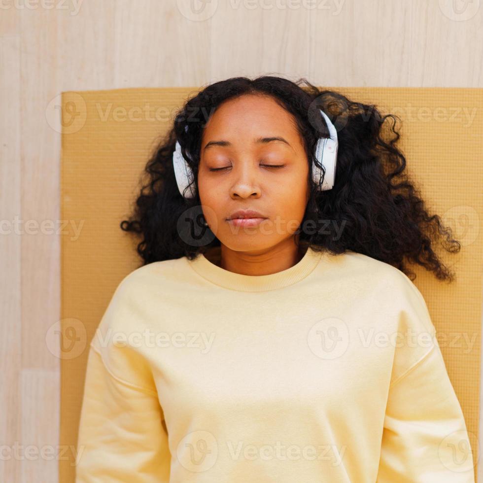 femme afro-américaine dans les écouteurs se reposant sur un tapis avec les yeux fermés. se détendre après l'exercice photo