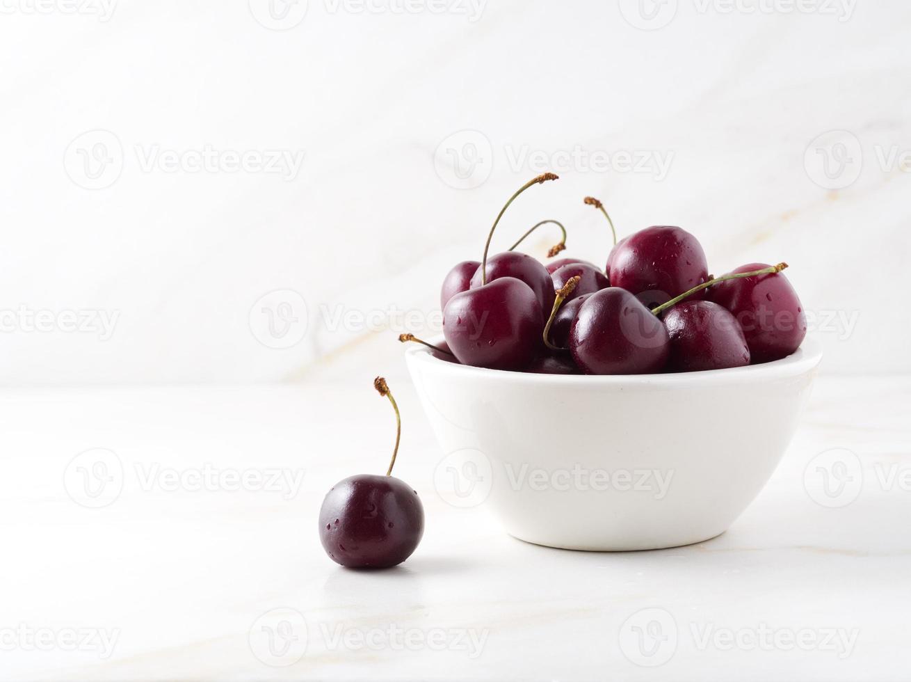 cerises douces rouges foncées dans un bol blanc sur une table blanche en pierre, vue latérale, espace pour copie photo