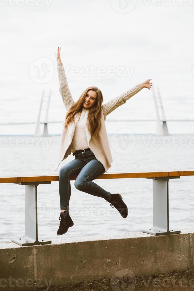 heureuse jeune fille assise sur le front de mer à la jetée du port, profitant de la vie et agitant les bras. femme aux cheveux longs sourit et profite du moment photo