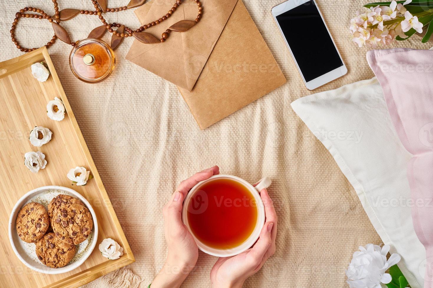 femme tenant une tasse de thé chaud. matinée confortable paresseuse au lit. accessoires femme à plat avec lettre, enveloppe, smartphone, parfum. photo