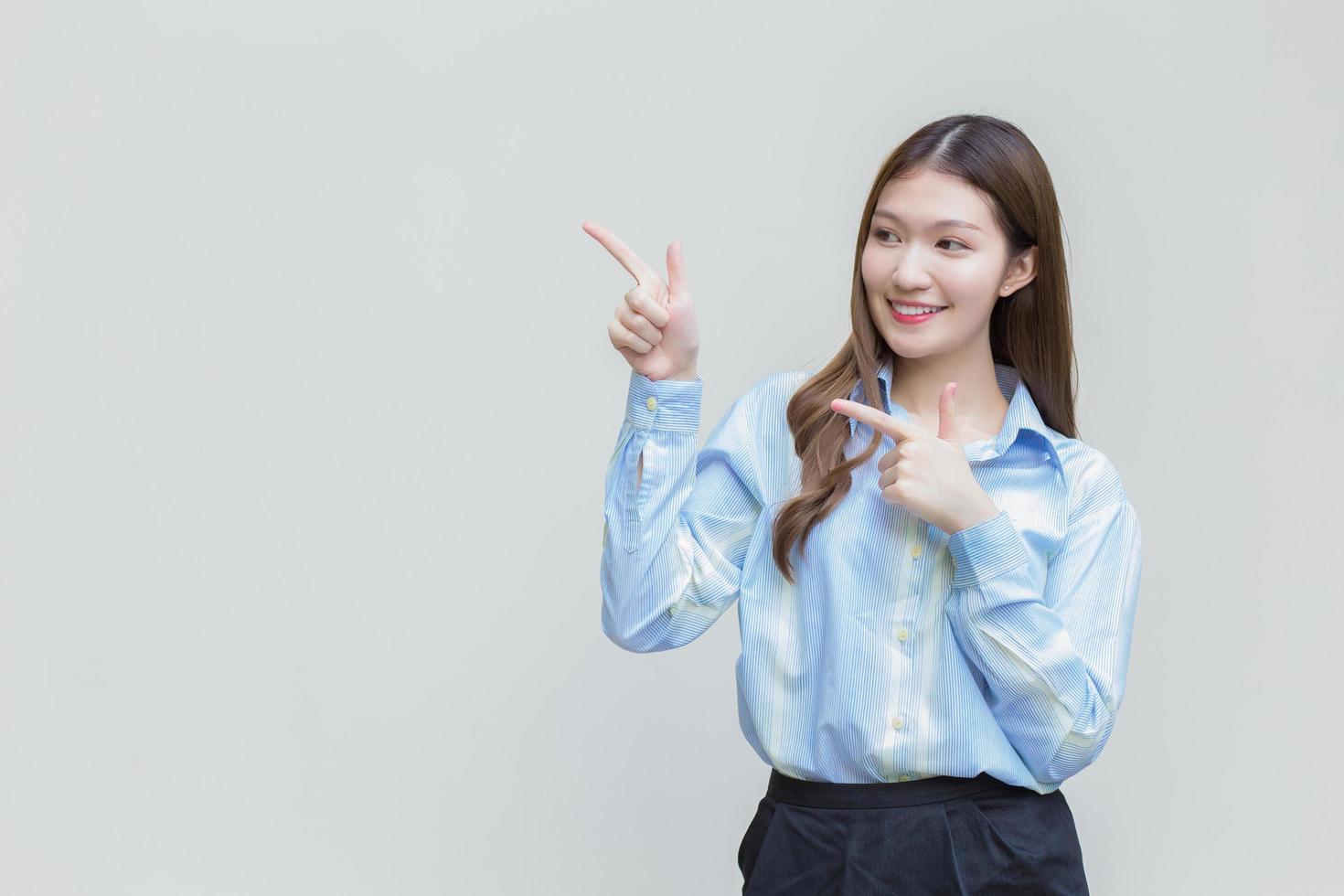 jeune femme d'affaires asiatique aux cheveux longs qui porte une chemise à manches longues bleue sourit joyeusement alors qu'elle montre le point vers le haut pour présenter quelque chose sur fond blanc. photo