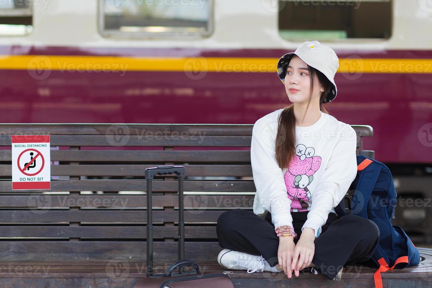 une femme asiatique en manches longues blanches et un chapeau est assis sur une chaise avec son sac sur la plate-forme du train dans la distanciation sociale comme un nouveau mode de vie normal. photo