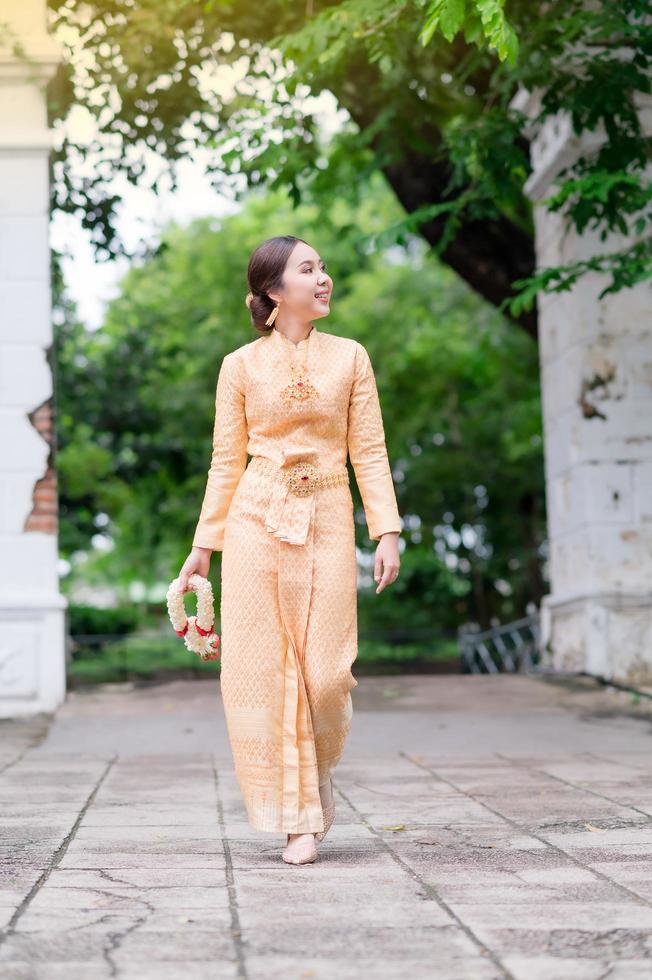 élégante femme thaïlandaise en robe thaïlandaise ornée de bijoux précieux se promène tenant des guirlandes de fleurs dans le temple thaïlandais photo