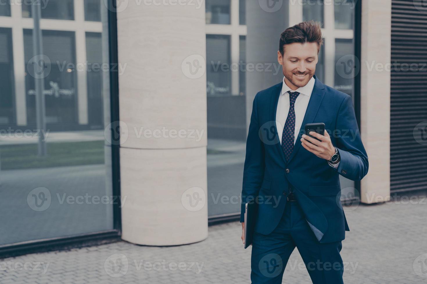 heureux entrepreneur masculin européen en costume tenant un téléphone portable et souriant à l'extérieur photo