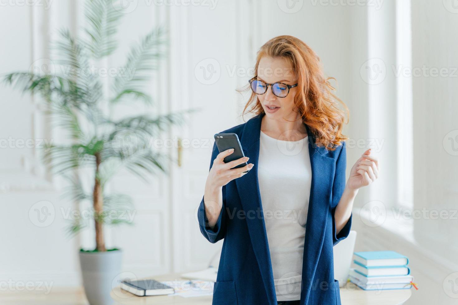 photo à la taille d'une femme d'affaires prospère utilise l'application sur un téléphone portable, lit les e-mails reçus du patron ou d'un collègue, vêtue de vêtements formels, pose à l'intérieur du bureau, découvre les nouvelles financières