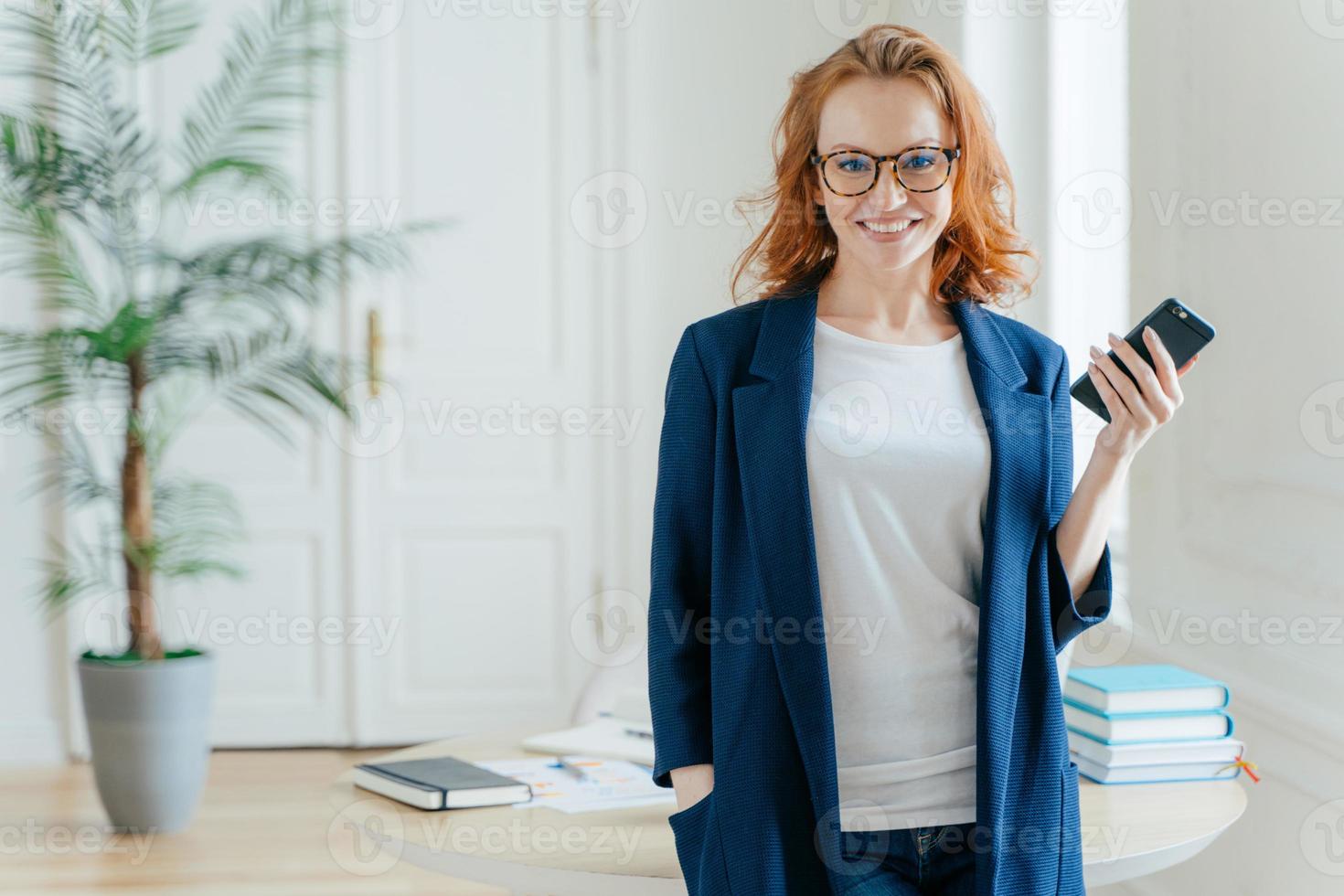 photo en demi-longueur d'une belle femme utilise un téléphone cellulaire moderne pour bloguer sur les réseaux sociaux, pose à l'intérieur du bureau, vêtue d'une tenue formelle, vérifie la notification, porte des lunettes optiques aux cheveux roux