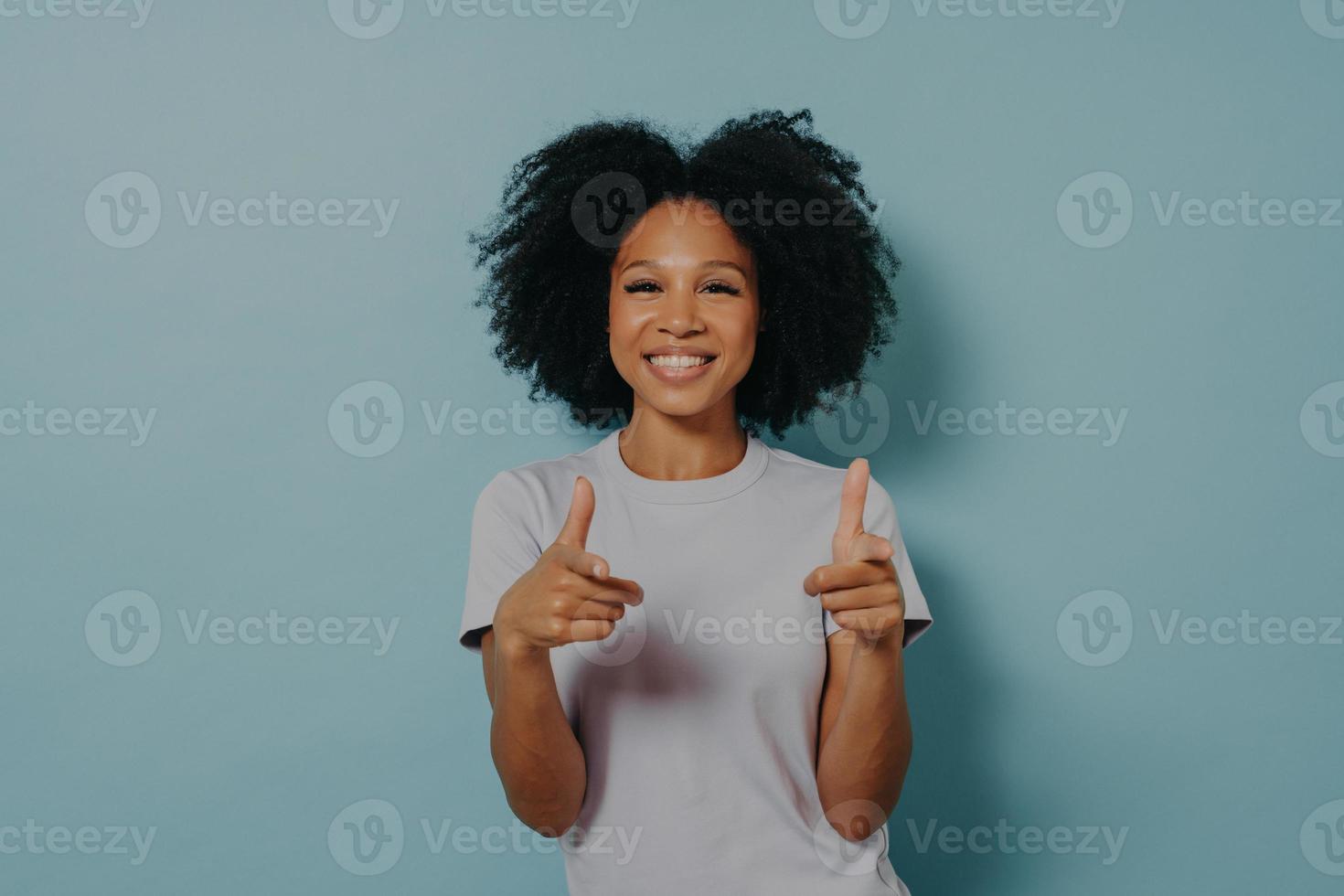 Dame à la peau sombre et amicale positive faisant un geste de pistolet à doigt en se tenant debout sur fond bleu photo