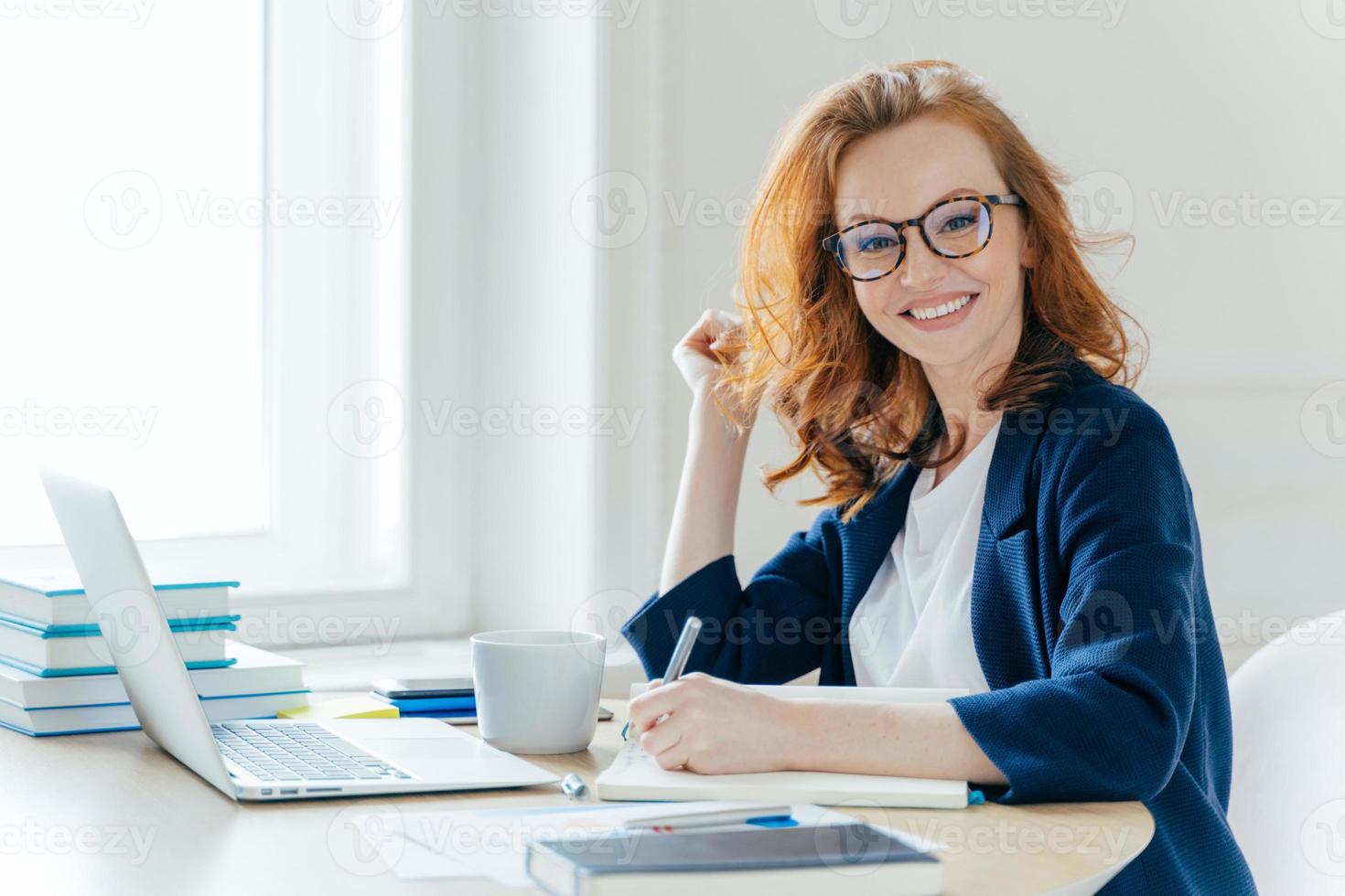 photo de côté d'une femme d'affaires prospère aux cheveux roux, sourit positivement, enregistre des informations dans le bloc-notes, boit du café, recherche des informations sur un ordinateur portable, développe un projet de démarrage