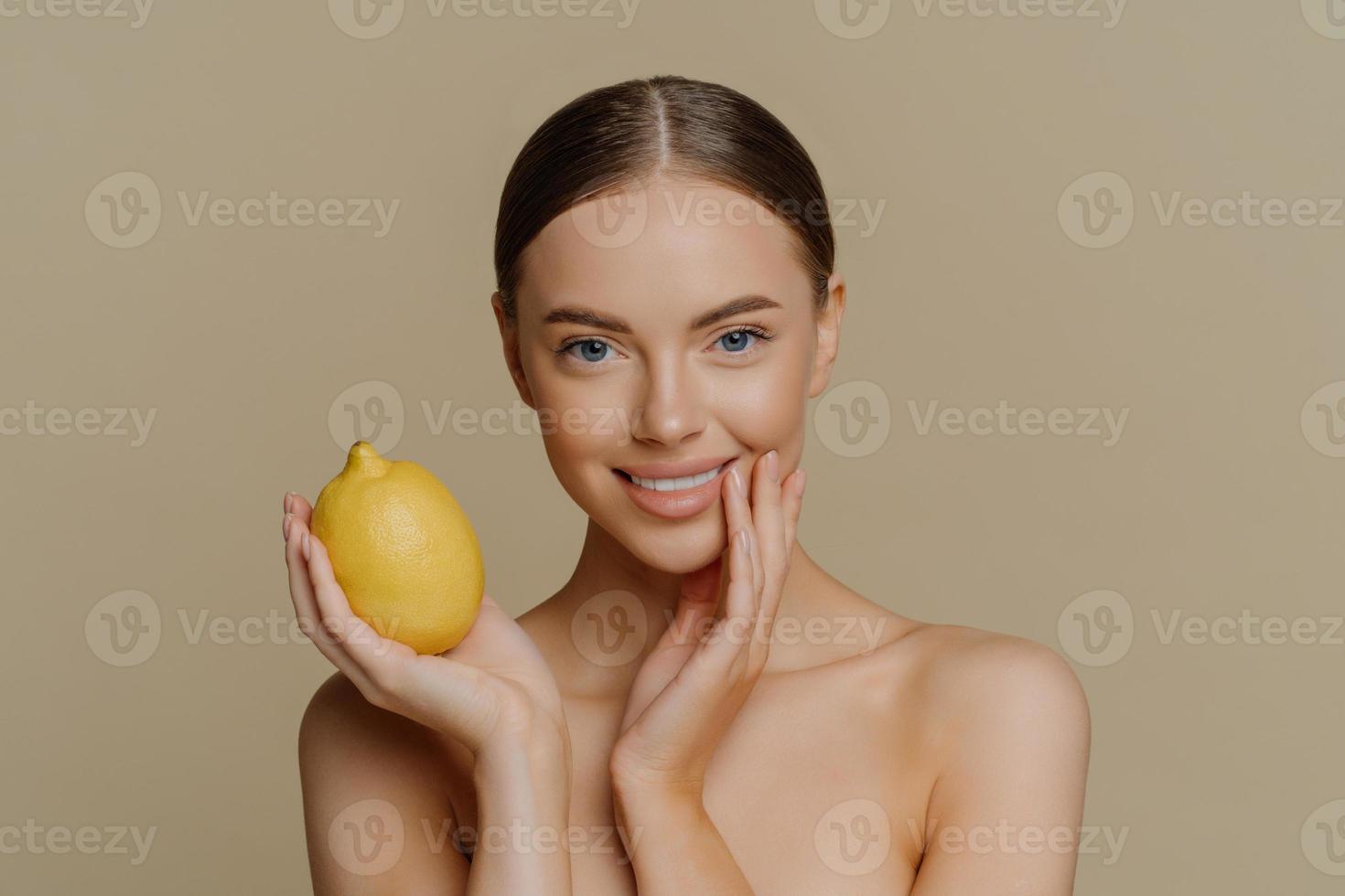 portrait d'une charmante femme à moitié nue aux cheveux noirs peignés contient du citron entier frais utilise des fruits pour les cosmétiques naturels pose sur fond marron avec un corps nu. concept de bien-être beauté des gens photo