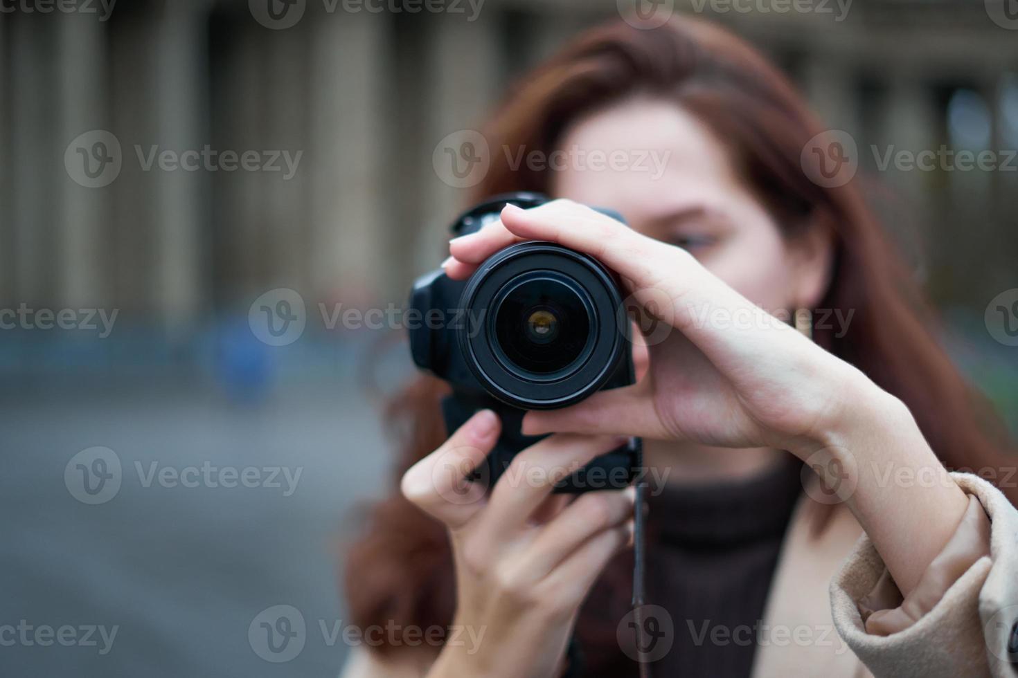 mise au point sélective sur l'objectif. belle fille à la mode élégante tient l'appareil photo dans ses mains et prend des photos. femme photographe aux longs cheveux noirs en ville, tournage urbain, vertical. personne méconnaissable