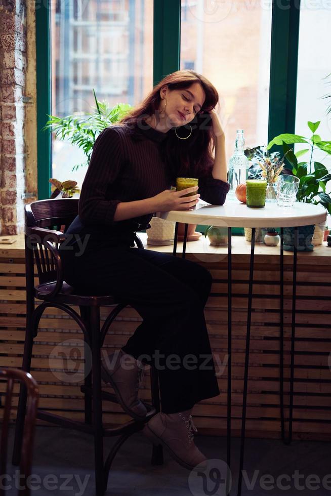 belle fille intelligente à la mode élégante et sérieuse est assise dans un café et boit un smoothie sain ou un latte végétalien. charmante femme réfléchie aux longs cheveux brun foncé. photo