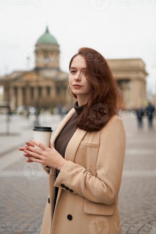 belle fille intelligente à la mode élégante et sérieuse tenant une tasse de café dans les mains va marcher dans la rue de st. Saint-Pétersbourg dans le centre-ville. charmante femme réfléchie aux longs cheveux noirs photo