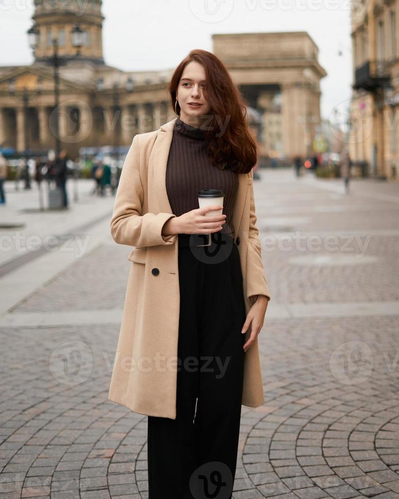 belle fille intelligente à la mode élégante et sérieuse tenant une tasse de café dans les mains va marcher dans la rue de st. Saint-Pétersbourg dans le centre-ville. charmante femme réfléchie aux longs cheveux noirs photo