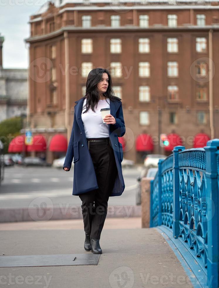 belle fille brune intelligente sérieuse tenant une tasse de café dans les mains va marcher dans la rue de st. Saint-Pétersbourg dans le centre-ville sur le pont. charmante femme réfléchie aux longs cheveux noirs erre seule photo