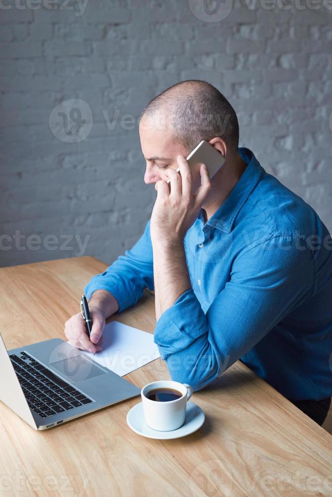 beau bel homme mûr écrit sur une feuille de papier et parle sur un téléphone portable, est assis devant un ordinateur, un ordinateur portable. homme avec des vêtements décontractés dans une chemise bleue à une table dans le bureau devant la fenêtre photo