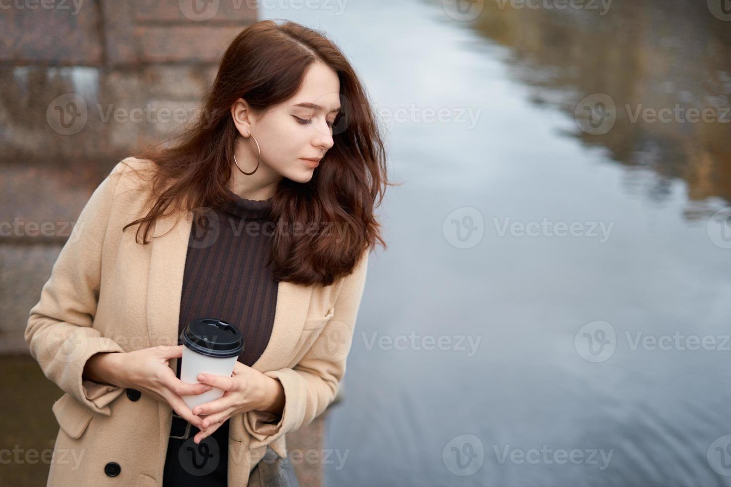 belle fille à la mode élégante et sérieuse tenant une tasse de café dans les mains marchant rue de st. Saint-Pétersbourg dans le centre-ville. charmante femme réfléchie avec de longs cheveux noirs regarde vers le bas, copiez l'espace photo