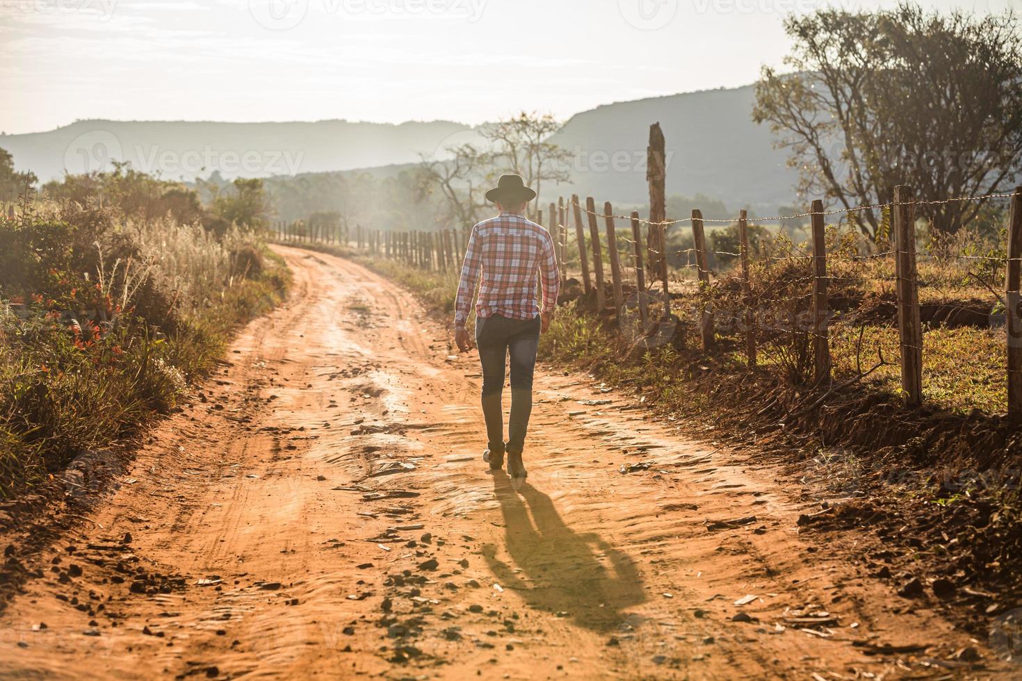 un travailleur masculin adulte portant un chapeau marche par une journée ensoleillée. ombre dure sur le chemin de terre rural. photo