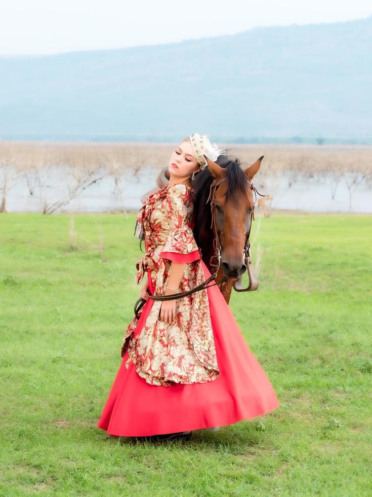 une belle femme occidentale dans une belle robe se tient avec son cheval dans la prairie du lac photo