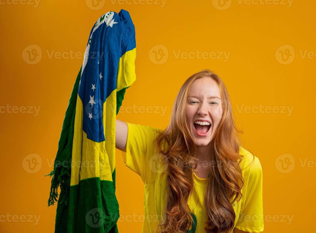 supporter du Brésil. fan de femme rousse brésilienne célébrant le football, match de football sur fond jaune. couleurs du Brésil. portant un t-shirt, un drapeau et un chapeau de fan. photo