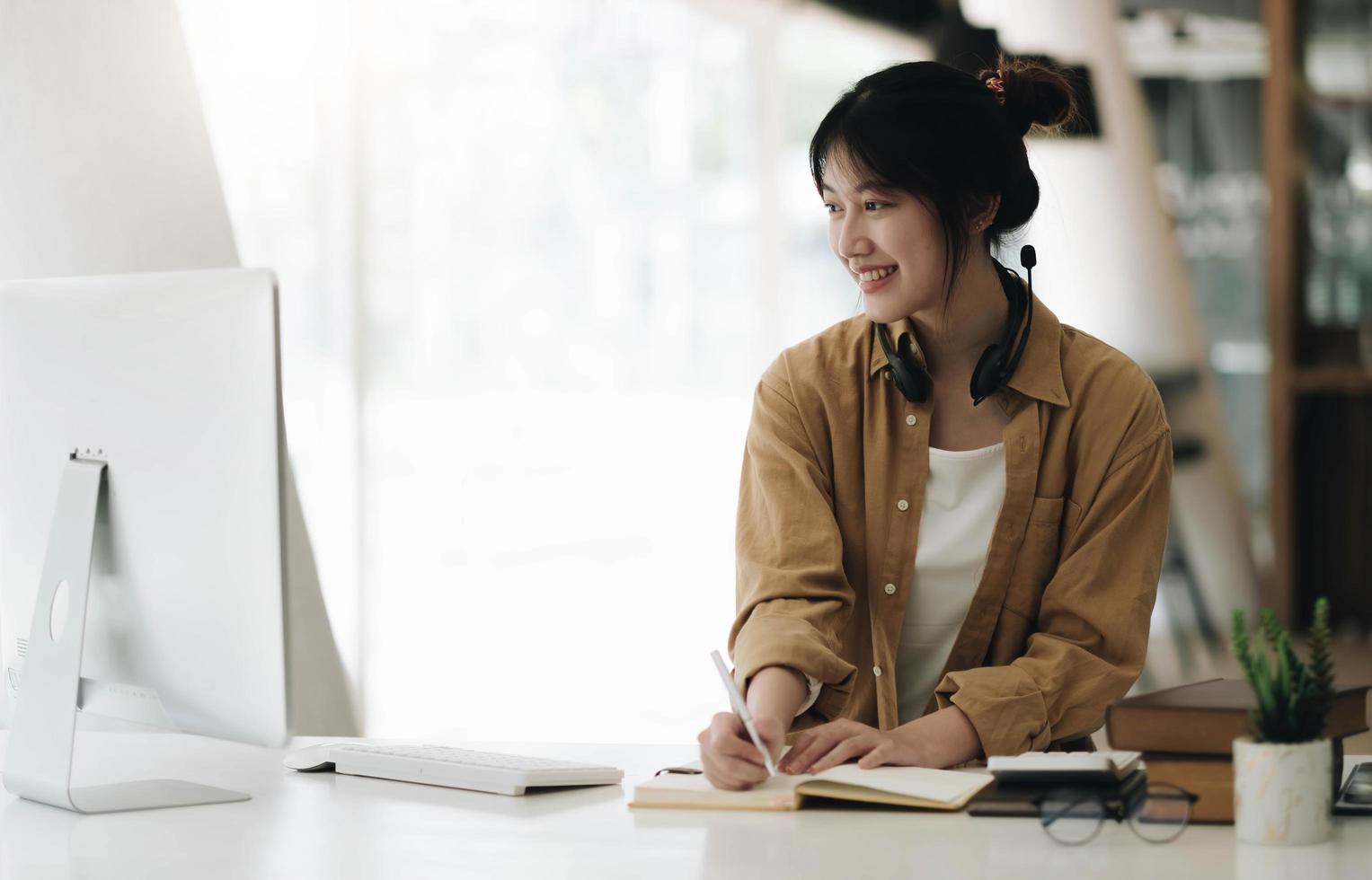 femme asiatique portant des écouteurs à l'aide d'un ordinateur portable et prenant des notes sur les devoirs que son professeur enseigne à la maison photo