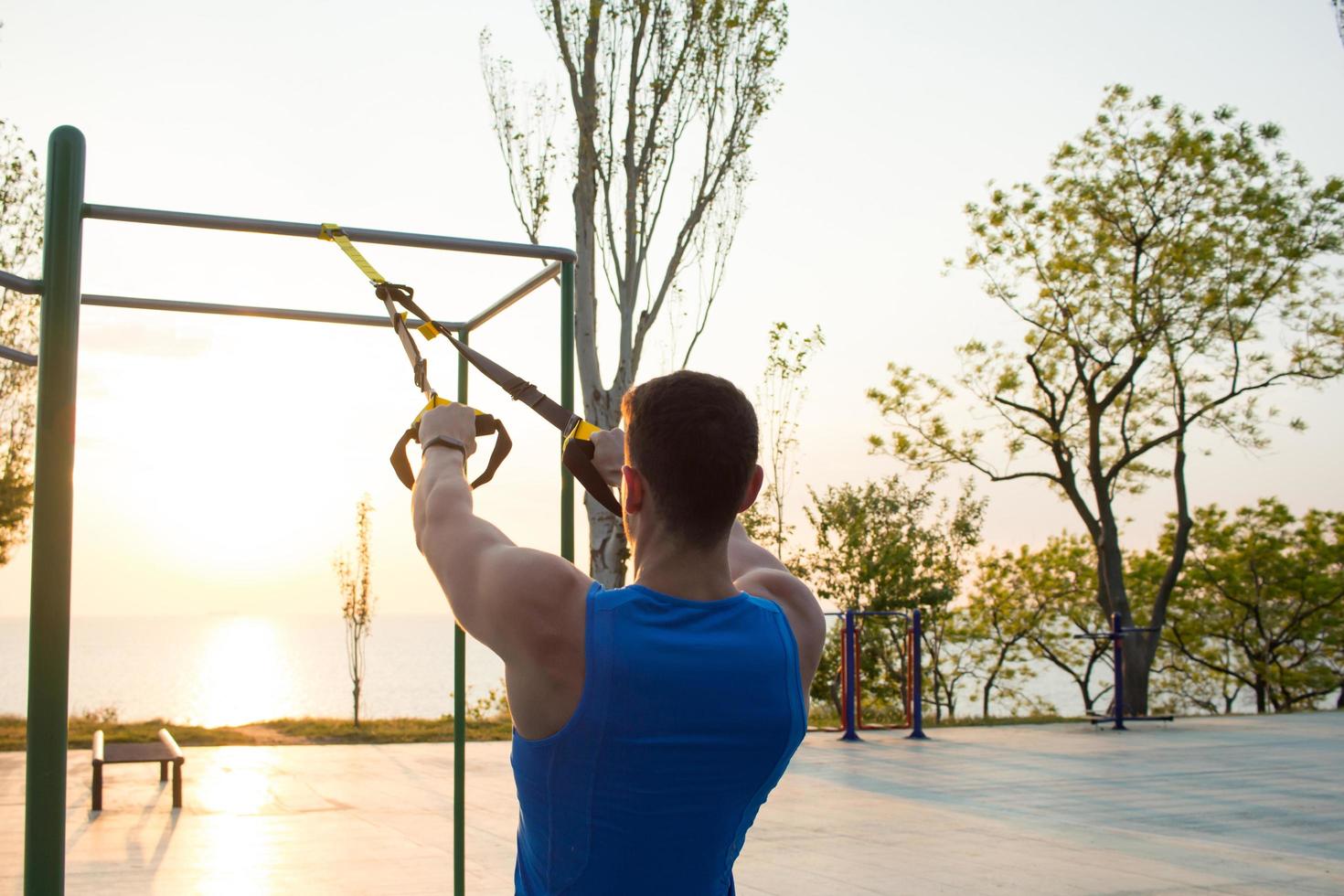 séance d'entraînement avec des sangles de suspension dans la salle de sport en plein air, homme fort s'entraînant tôt le matin sur le parc, lever ou coucher de soleil sur fond de mer photo