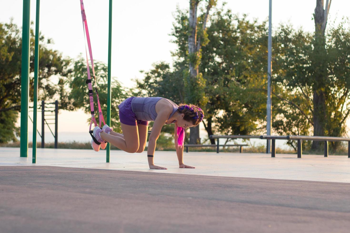 belle femme en forme de sportwear rose et violet s'entraînant sur une salle de sport en plein air le matin, exercices avec des sangles de suspension dans le parc photo